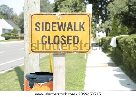 sidewalk closed writing text orange black horizontal rectangle sign on wood post and grass next to street road and sidewalk