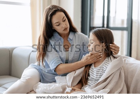 Mother worries about her sick daughter, touching lymph nodes on neck while sitting on sofa near. Girl kid wrapped in plaid feeling weakness, fever and respiratory infection symptoms Royalty-Free Stock Photo #2369652491