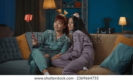 Medium-full photo capturing two young women in pajamas sitting on the couch and taking photos on a smartphone using a selfie stick, wearing funny headbands.