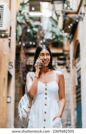 Latin American tourist with backpack holding hand in pocket of white dress and looking away with smile while talking on mobile phone on blurred background of narrow street in Barcelona, Spain Royalty-Free Stock Photo #2369682505