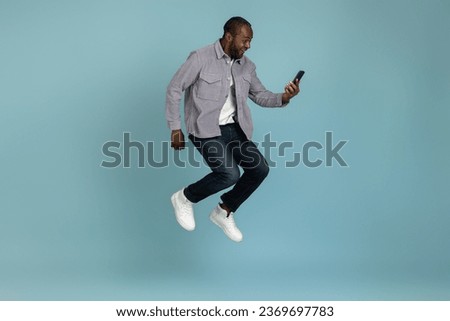 Overjoyed black man wearing gray shirt using smartphone jumping up Royalty-Free Stock Photo #2369697783