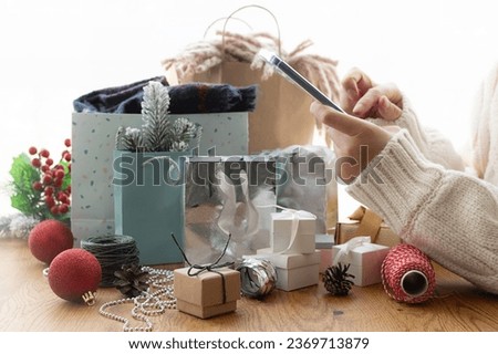 Woman’s hands with smartphone and Christmas gifts and decorations on the table close up. Festive internet shopping concept. Buy online composition.