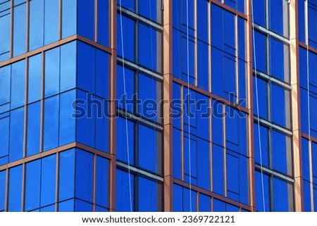 A modern and abstract architectural photo of blue glass windows reflecting white clouds and blue sky. The windows are framed by red metal lines that create a contrast and a geometric pattern. 