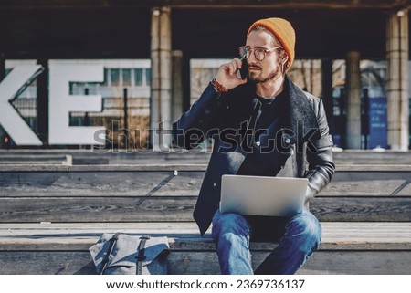 millennial man sitting with netbook and talking via cellphone Royalty-Free Stock Photo #2369736137