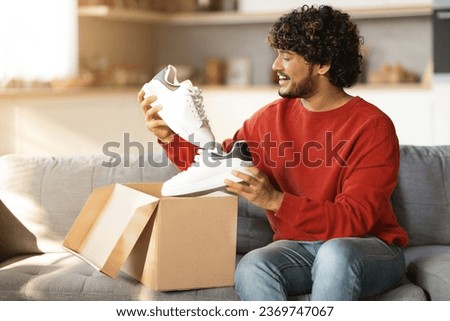 Shopping And Delivery. Cheerful Indian Customer Guy Holding Shoes Unpacking Cardboard Box With Delivered Footwear While Sitting On Couch At Home. Commerce And Retail Concept, Free Space Royalty-Free Stock Photo #2369747067