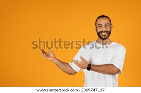 Cheerful adult european man with beard in white t-shirt points with hands to copy space, isolated on orange studio background. Recommendation sale, ad and offer, presentation