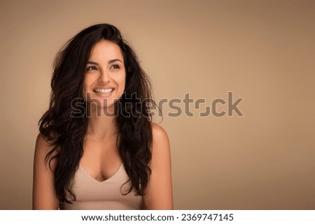 Cheerful beautiful millennial eastern lady with long gorgeous hair and silky glowing youthful skin posing on beige studio background, looking at copy space and smiling. Hair and face care concept Royalty-Free Stock Photo #2369747145
