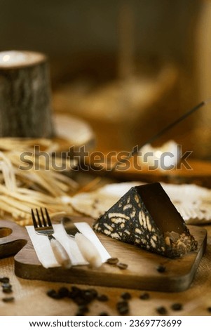 A slice of mosaic cake presented on burlap, alongside wooden cutlery and board. Captured with incense and warm candlelight. Royalty-Free Stock Photo #2369773699