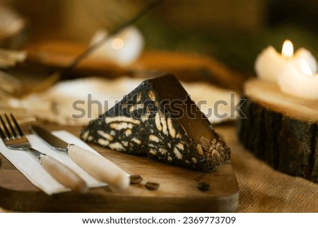 A slice of mosaic cake presented on burlap, alongside wooden cutlery and board. Captured with incense and warm candlelight. Royalty-Free Stock Photo #2369773709
