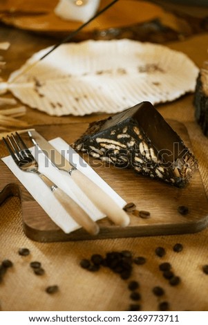 A slice of mosaic cake presented on burlap, alongside wooden cutlery and board. Captured with incense and warm candlelight. Royalty-Free Stock Photo #2369773711
