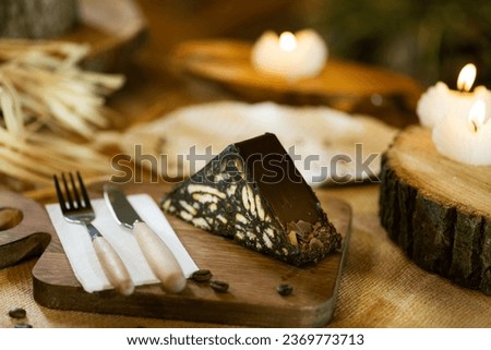 A slice of mosaic cake presented on burlap, alongside wooden cutlery and board. Captured with incense and warm candlelight. Royalty-Free Stock Photo #2369773713
