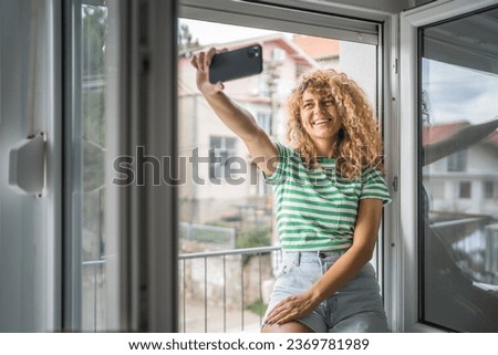 One woman adult caucasian female stand at window at home in bedroom use mobile phone hold smartphone take selfie photos self portrait or make a video call daily morning routine happy smile copy space
