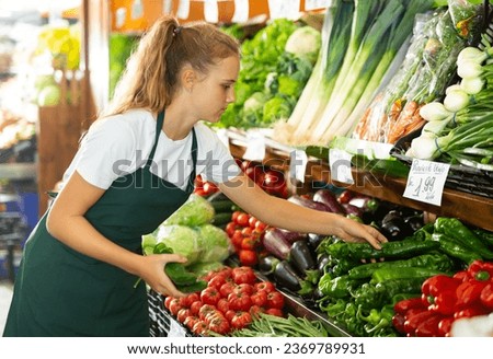 Focused fifteen-year-old girl who works part-time in a store as a trainee saleswoman puts green pepper on showcase Royalty-Free Stock Photo #2369789931