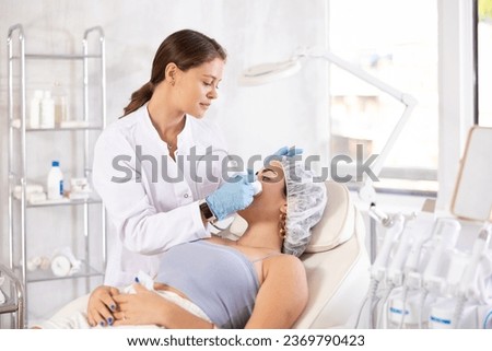 European female aesthetician making facial massage with vibration attachement of beauty machine to young girl Royalty-Free Stock Photo #2369790423