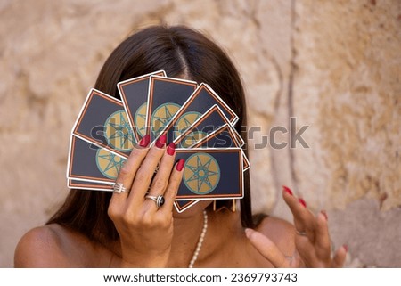 A young dark-haired girl wearing gold earrings and a pearl necklace around her neck smiles enigmatically and covers her eyes with a fan of tarot cards. Young brunette fortune teller holding tarot 