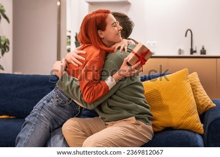 Man woman hugging sitting on couch at home celebrating anniversary, giving gift. Happy affectionate wife cuddling husband with smiling happy face, present in hand. Family celebration, feast concept. Royalty-Free Stock Photo #2369812067