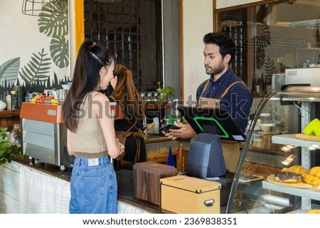 Young Indian barista or cashier worker man hold payment machine accept credit card cashless transaction from Asian customer woman, modern technology paying consumer eatery product cafe and bakery shop