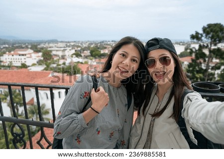 two smiling asian chinese female friends looking at camera together while taking selfie picture with beautiful clock tower view backdrop on balcony of santa Barbara county courthouse in California