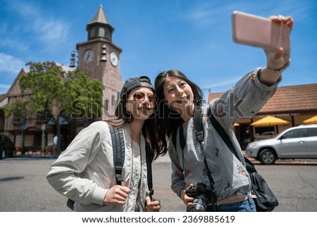 two smiling asian chinese sister tourists holding smartphone and taking photo together with Danish style brick clock tower in the back against blue sky in solvang California usa