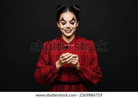 Young woman with Halloween makeup face art mask wear clown costume red dress hold in hand use mobile cell phone isolated on plain solid black background studio portrait. Scary holiday party concept