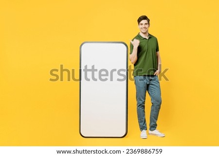 Full body side view young happy man wears green t-shirt casual clothes point thumb finger on big huge blank screen mobile cell phone smartphone with workspace area isolated on plain yellow background