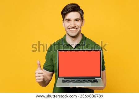 Young happy IT man he wearing green t-shirt casual clothes holding use work on laptop pc computer with blank screen workspace area show thumb up isolated on plain yellow background. Lifestyle concept