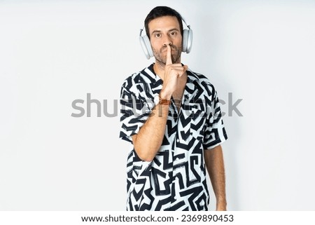 young handsome businessman wearing printed shirt making hush gesture with finger on her lips wearing  wireless headphones. Be quiet. Royalty-Free Stock Photo #2369890453