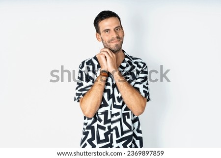 Charming serious young handsome man wearing printed shirt keeps hands near face smiles tenderly at camera