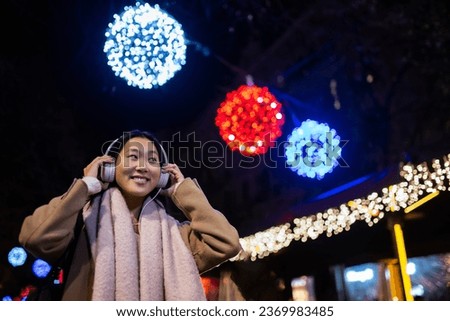 Young happy woman wearing headphones with christmas lights behind. Joyful girl listening to music at the street during winter.