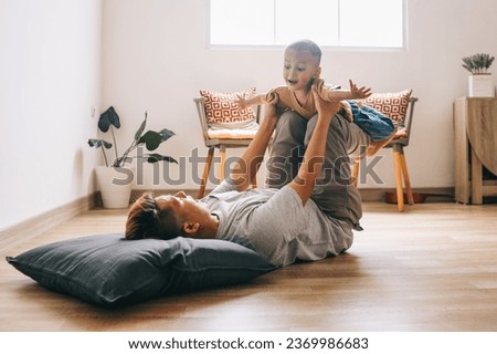 Joyful young Asian father lying on parquet floor and playing plane fly with son  Royalty-Free Stock Photo #2369986683