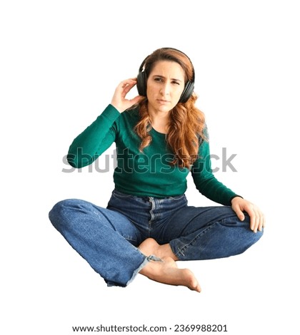 Unhappy young brunette woman sitting on the floor while wearing headphones against a white background