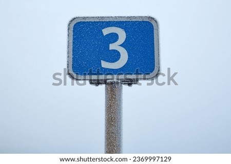 A road sign covered in frost stands on the roadside, surrounded by a misty winter landscape.