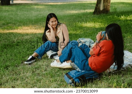 Young asian woman taking picture of her smiling friend with instax camera while sitting on grass in green park