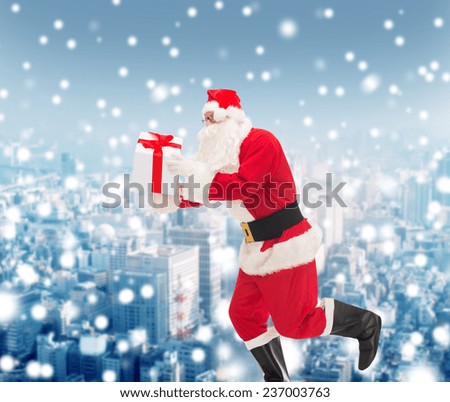 christmas, holidays and people concept - man in costume of santa claus running with gift box over snowy city background