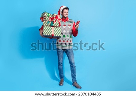 Full length photo of nice young male hold device eshop presents pile dressed x-mas print vest hat garment isolated on blue color background