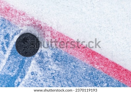 Looking down on a black ice hockey puck plowing through snow and stopping on the edge of the Goal Line of the goal crease. 