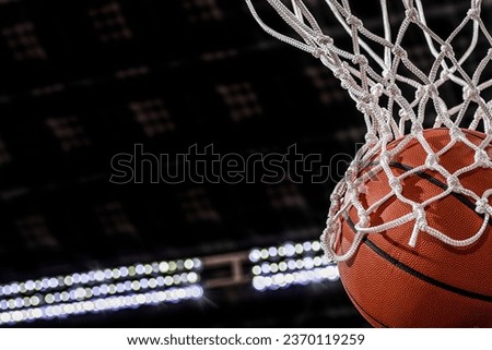 A close-up view of an orange basketball falling through the rim and a white nylon net with the arena lights in the background.