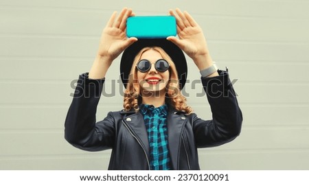 Portrait of stylish happy laughing woman stretching her hands for taking selfie with mobile phone wearing black round hat, leather jacket and sunglasses on gray background