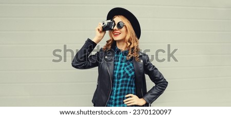 Portrait of beautiful young woman photographer with film camera taking picture wearing black round hat, leather jacket on gray background