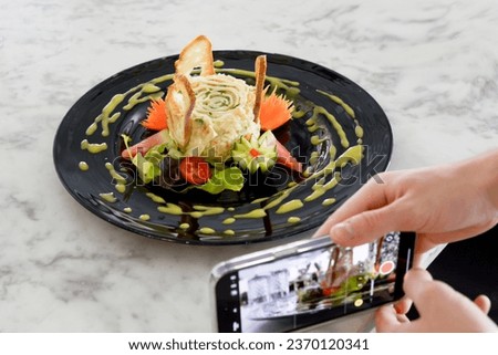 hand holding phone,taking a photo of Olivier salad with mayonnaise shaped into a cake,decorated with beautifully chopped vegetables on a plate with a decorative pattern of spices,gray sauce,selective