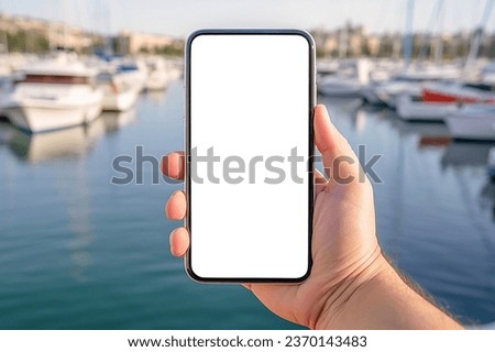 Businessman holding smartphone with blurred yacht marine background. White screen, mockup. People tourism travel and spare time concept. modern mobile phone with blank screen with harbor background
