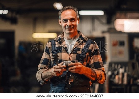 A portrait of a grey haired machinist showing a metal cylinder in a factory, workshop setting. He has calipers in his hands and the machine part. His protective glasses are hanging on his overalls. Royalty-Free Stock Photo #2370144087