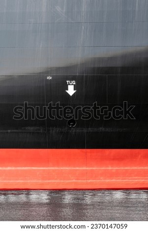 Section of the black and red hull of a ship with letters TUG and arrow pointing to waterline and location to secure tow of tugboat