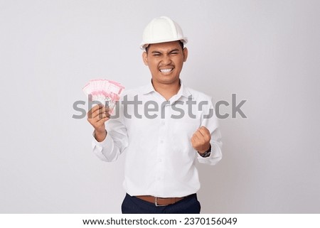 Excited young Asian professional engineer employee man in a protective helmet hardhat holding money and celebrating success isolated on white background