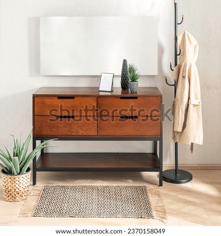 Mid-Century Modern Wooden Chest of Drawers Dresser and Metal Coat Rack in a Bright Living Room with a Blank Mock-up Poster. A Beige Women Jacket Hanging on the Coat Rack, Decorative Potted Plants. Royalty-Free Stock Photo #2370158049