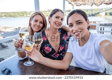 Three cheerful girlfriends sit at a table with a glass of wine drinking, talking, and taking a selfie on a restaurant terrace by the river.