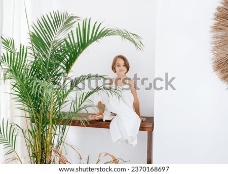 A girl in a white summer dress sits on a wooden table at home behind a tropical palm tree