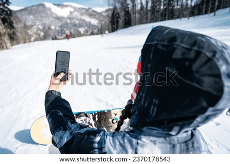 Unrecognizable female snowboarder sitting on snow and taking a break while using smartphone to make a photo or have a video call. Unrecognizable person taking a selfie on a ski track.