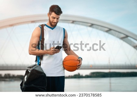 Young tall sportsman walking with sports bag while using mobile phone and holding a basketball. Handsome male basketball player with ball and sports bag holding and looking at mobile phone.