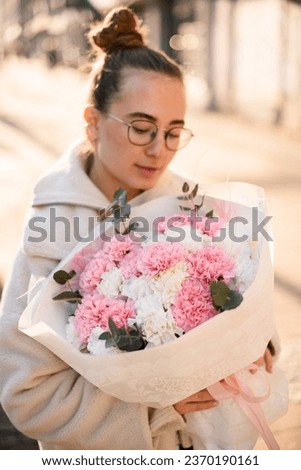 Young woman holding freshly made minimalistic blossoming flower bouquet of pink, white carnations decorated with eucalyptus branches and pink feathers. Blurred background